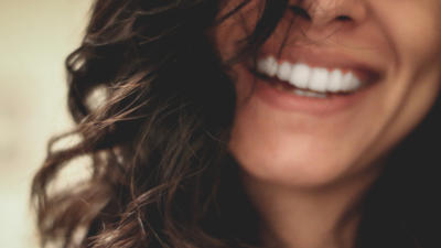 Close-up, slightly out of focus image of woman's head from nose to chin, smiling with white teeth and brown hair slightly obstructing face